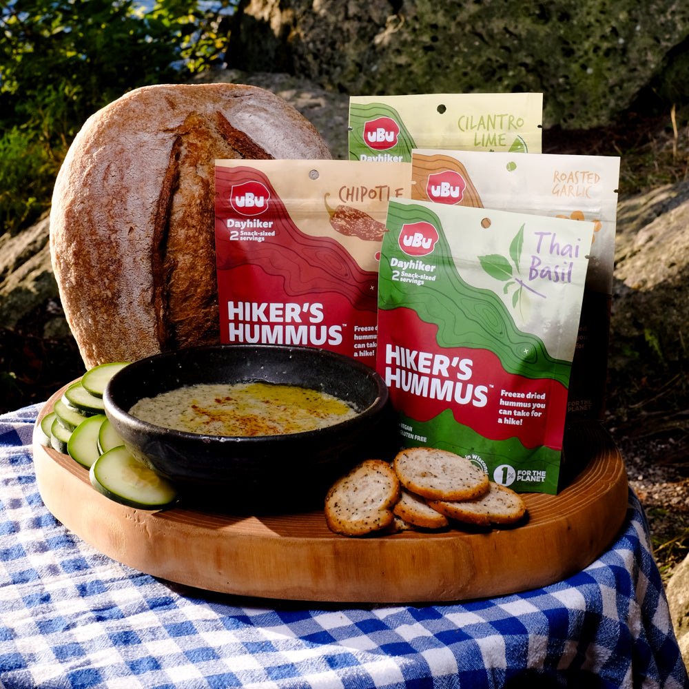 The four flavors of uBu's Hiker's Hummus are situated on a wooden cutting board, next to a loaf of delicious sourdough bread, cucumbers, pita chips, and a bowl of the hummus reconstituted. The cutting board is on a blue check tablecloth on what appears to be a stump. All of this is situated against a boulder and various bits of foliage. 